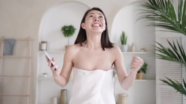 Opgewonden multiculturele aziatische meisje met smart phone schreeuwen ja winnende online spel, vieren bod te winnen overwinning, het krijgen van e-commerce winkelen aanbod, tijdens lichaamsverzorging en schoonheid procedure in de badkamer — Stockvideo