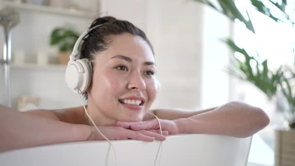 Atractiva joven sensual mujer morena asiática con auriculares disfrutando de un baño caliente. Chica milenaria multiétnica escuchando música chill relajante en el baño, terapia de tratamiento, concepto de estilo de vida saludable — Vídeo de stock