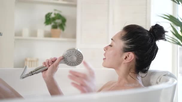 Emocionada mujer asiática multicultural cantando en bañera usando cabezal de ducha. Felices mujeres milenarias divirtiéndose disfrutan del cuidado corporal diario en el baño, terapia de tratamiento de belleza y concepto de estilo de vida saludable — Vídeo de stock