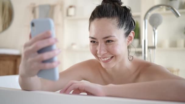 Mujeres multiculturales felices haciendo videollamadas usando un smartphone sentado en la bañera. Millennial chica asiática con teléfono móvil teniendo webcam vídeo conversación disfrutar de diario bodycare en cuarto de baño — Vídeo de stock