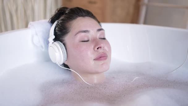 O tânără asiatică atractivă cu căști cântând în cadă cu ochii închiși. Fata multi-etnică milenară ascultând muzică rece relaxare în baie, terapie de îngrijire a corpului, concept de stil de viață sănătos — Videoclip de stoc