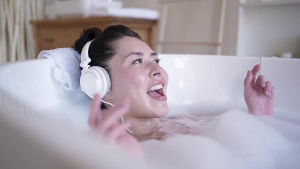 Attraktiv ung asiatisk kvinna med hörlurar som sjunger i badkar med slutna ögon. Multi etnisk tusenårig dam lyssnar chill musik avkopplande i badrummet, behandling terapi, hälsosam livsstil koncept — Stockvideo