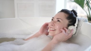 Küvette gözleri kapalı şarkı söyleyen kulaklıklı çekici Asyalı bir kadın. Çok ırklı milenyum kızı rahat müzik dinliyor banyoda dinleniyor, tedavi görüyor, sağlıklı yaşam tarzı kavramları var.