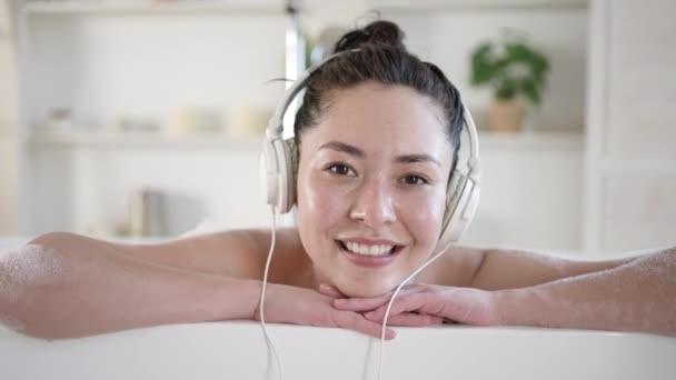 迷人而快乐的年轻混血亚洲女人，带着耳机在浴缸里歌唱。多民族千禧年女孩在浴室听清凉的音乐放松，治疗疗法健康的生活方式理念 — 图库视频影像