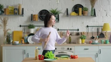 Genç ve mutlu melez ırk kızı elinde mikrofonla şarkı söylüyor mutfakta müzik dinliyor, komik Asyalı kadın sebze salatası hazırlıyor sağlıklı yemekler pişiriyor evde tek başına eğleniyor.