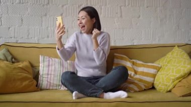 Şaşırtıcı mutlu Asyalı kız evde akıllı telefondan ecommerce alışveriş teklifi almasını kutluyor. Heyecanlı karışık yarış müşterisi cep telefonuna bakarak mobil zafer kavramını kutlayan bir uygulama kullanıyor.