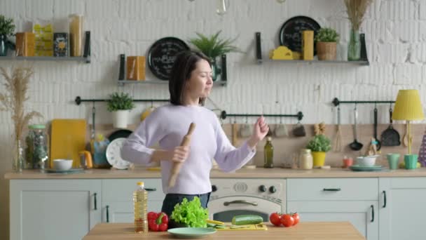 Feliz chica de raza mixta joven sosteniendo micrófono batidor cantando canción bailando escuchando música en la cocina, divertida mujer asiática preparando ensalada de verduras cocinando comida saludable divirtiéndose sola en casa — Vídeos de Stock