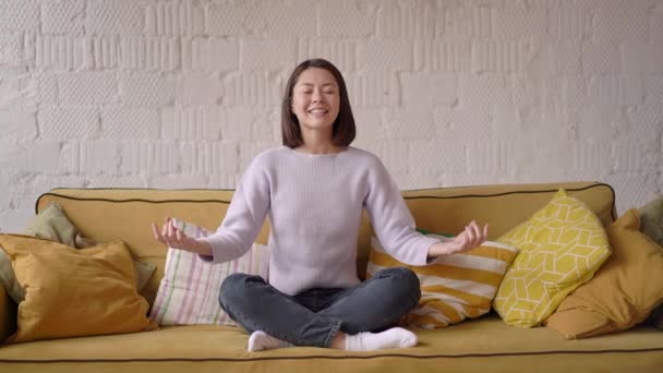 Mindful calma hermosa mujer de raza mixta sentarse en el sofá haciendo yoga respiración ejercicio en casa tomar descanso relajarse ojos cerrados, joven sonriente chica asiática meditar en casa sentirse zen sin estrés paz concepto de la mente — Vídeos de Stock