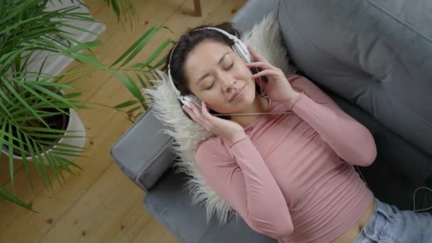 Jovem mulher asiática relaxante no sofá confortável com os olhos fechados usando fones de ouvido, milenar senhora gosta de ouvir música chill e som de áudio meditando, relaxando em casa. Visão superior em câmera lenta — Vídeo de Stock