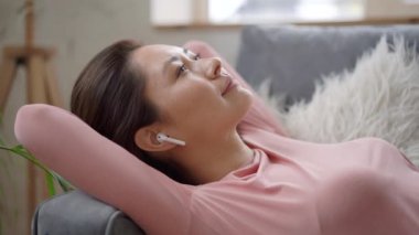 Karışık ırk genç Asyalı kadın rahat koltukta dinleniyor gözleri kablosuz kulaklıkla kapalı. Güzel bayan evde meditasyon yaparken ve hayal kurarken dinlenmeyi seviyor.
