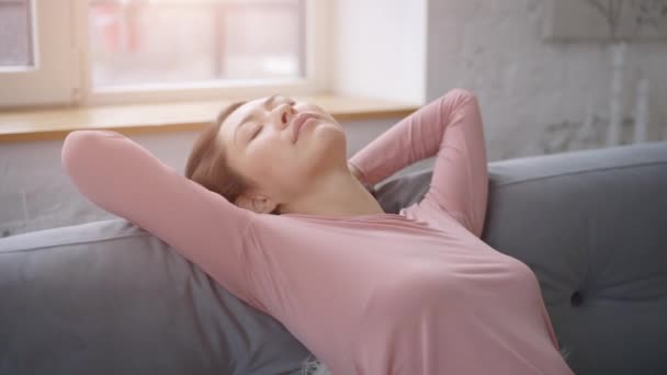 Attraktive junge asiatische Frau, die auf dem gemütlichen Sofa ruht, tief Luft schnuppert und die Hände hinter dem Kopf hält, fröhliche ruhige ethnische Dame, die sich entspannt und auf der bequemen Couch träumt, fühlt sich stressfrei an — Stockvideo