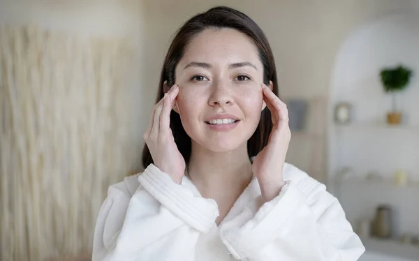 Vonzó többnemzetiségű hölgy néz tükörbe, megérintve az egészséges puha arcbőrt a fürdőszobában. Fiatal vegyes fajú ázsiai nő csinál masszázs elégedett arc eljárások bőrápolás koncepció. — Stock Fotó