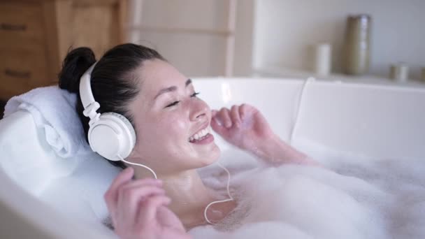 Atractiva joven asiática hembra con auriculares cantando en bañera con los ojos cerrados. Multi étnica milenaria dama escuchando música chill relajante en el baño, terapia de tratamiento, concepto de estilo de vida saludable — Vídeo de stock