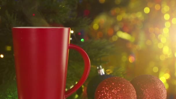 Red cup of steaming coffee next to a decorated Christmas tree — Stock Video