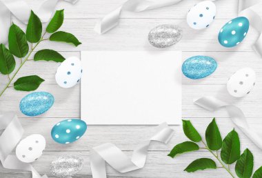 Gümüş, beyaz ve mavi yumurtalardan, kurdelelerden, ağaç yapraklarından, beyaz ahşap arka planda bir kağıt yaprağından oluşan paskalya kompozisyonu. Paskalya tatili için bahar reklamı. Üst görünüm, kapat, boşluğu kopyala