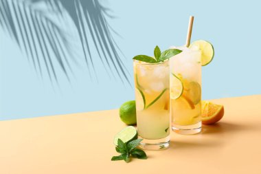Mavi ve bej arka planda portakal ve limon dilimli iki soğuk mojito kokteyli. Güneşli gölgeli serinletici yaz içeceği.