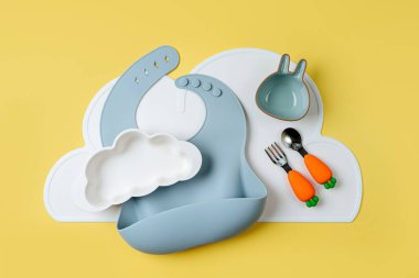 Sarı arka planda bulut şeklinde bebek önlükleri ve tabaklar. Bebeğe servis yapıyorum. Çocuk menüsü, beslenme ve beslenme kavramı 