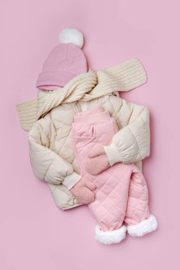 Kabarık ceket, kalın pantolon, pembe arka planda şapka ve eşarp. Kış için bebek kıyafetleri. Moda çocuk kıyafeti..
