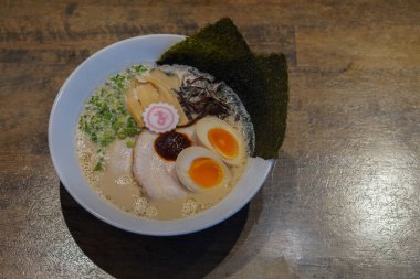Donkatsu ramen, yarım yumurta, yosun, dilimlenmiş domuz eti, acı biber ezmesi, sebze, ahşap bir zemine yerleştirilmiş beyaz seramik bir kase.