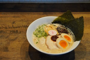 Donkatsu ramen, yarım yumurta, yosun, dilimlenmiş domuz eti, acı biber ezmesi, sebze, ahşap bir zemine yerleştirilmiş beyaz seramik bir kase.