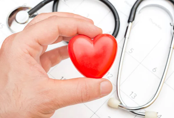 Man\'s hand holding red heart over calendar and stethoscope.