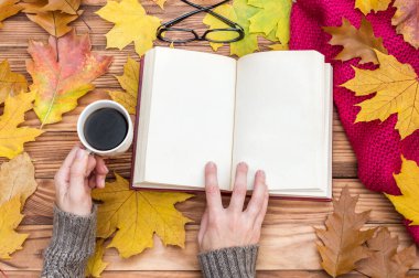Masada sonbahar yaprakları, bir fincan kahve ve örgü atkıyla kitap okuyan bir kadın. Metin için boşluk.