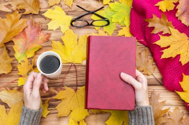 Masada sonbahar yaprakları, bir fincan kahve ve örgü atkıyla kitap okuyan bir kadın. Metin için boşluk.