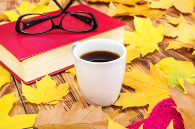 Kitaplı bir fincan kahve, örgü eşarp ve sonbahar yaprakları masanın üzerinde..