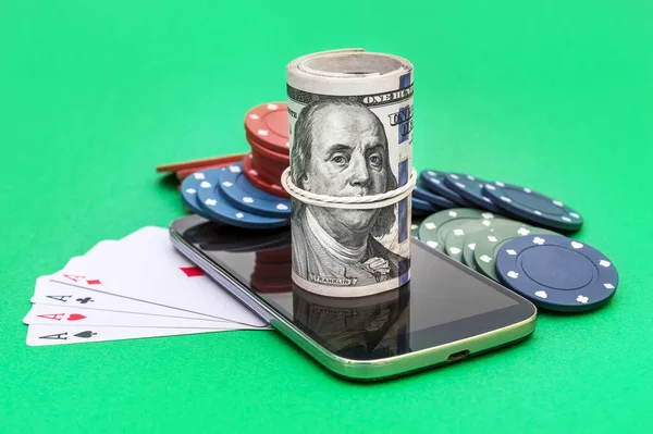Smartphone with blank screen, poker chips, playing cards and money on green background.