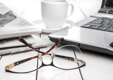 Eyeglasses with newspaper, laptop and cup of coffee on the white table.
