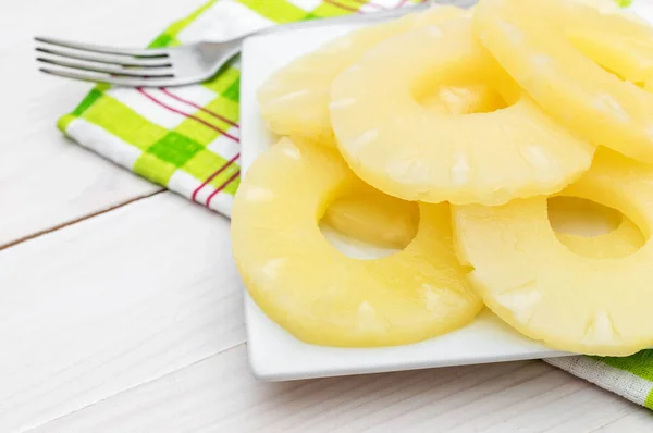 Plate with canned pineapple rings with fork and kitchen towel on white wooden table.