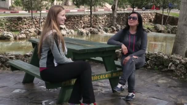 Dos hermosas mujeres hablando entre ellas, sentadas en un banco del parque — Vídeo de stock