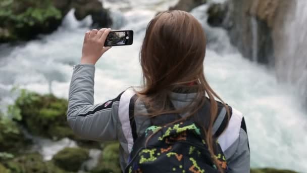 Közeli kép: egy fiatal lány, állva a hátizsákjába veszi a smartphone hegyi vízesés — Stock videók