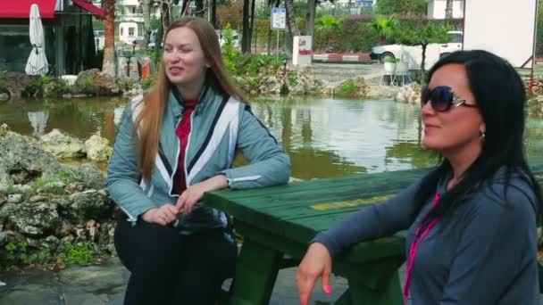 Duas jovens mulheres alegres sentadas em um banco conversando em uma mesa no parque — Vídeo de Stock