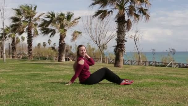 Joven mujer hermosa sentada en la hierba verde en el parque, contra el telón de fondo de las palmeras . — Vídeo de stock