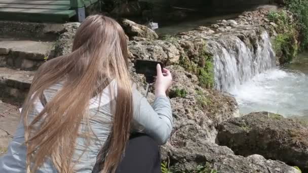 Junge Frau beim Fotografieren von Wasserfällen. junge Frau mit Smartphone macht das Foto vom Wasserfall an einem sonnigen Tag im Park. — Stockvideo