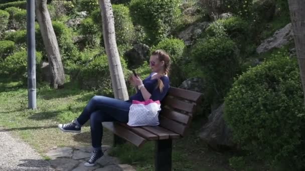 Hermosa joven feliz mirando la pantalla del teléfono inteligente sentado en un banco en el parque en el verano — Vídeo de stock