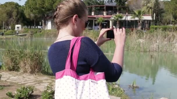 夏の公園では、太陽の光でスマート フォンを使用して写真を作る美しい幸せな若い女の子 — ストック動画