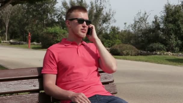 Joven alegre hombre feliz hablando con el teléfono inteligente sentado en un banco en el parque en el verano — Vídeo de stock