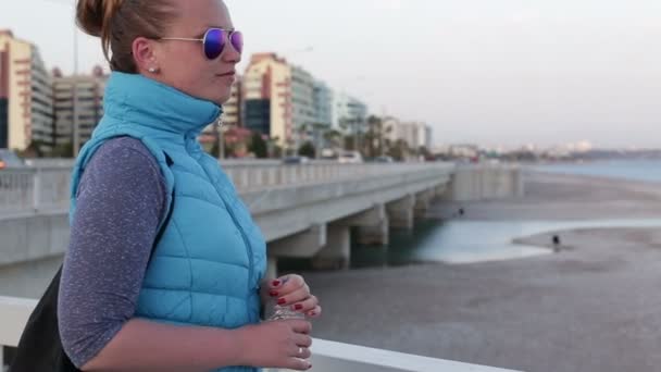 Hermosa chica de pie en el paseo marítimo sosteniendo una botella de agua — Vídeos de Stock