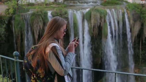 Schöne fröhliche fröhliche junge blonde Mädchen schreibt eine Nachricht, um ein Smartphone vor dem Hintergrund eines großen Wasserfalls verwenden — Stockvideo