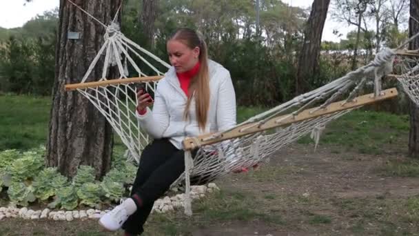 Krásná mladá dívka píše zprávu pomocí smartphone seděla v houpací síti s úsměvem — Stock video