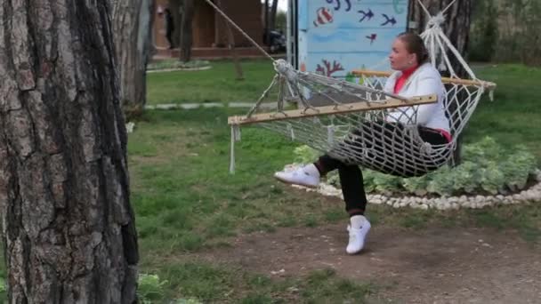 Beautiful young girl talking to use a smartphone limbs into a hammock and sitting in hammock smiling — Stock Video