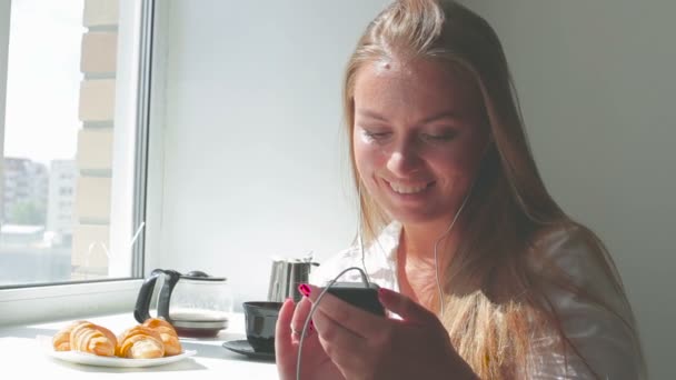 Jovem menina bonita tem café da manhã ouvir música usar smartphone em um dia ensolarado — Vídeo de Stock