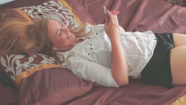 Όμορφη νεαρή κοπέλα ξαπλωμένη στο κρεβάτι και χρήση smartphone χαμογελώντας — Αρχείο Βίντεο