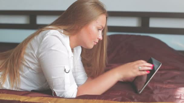 Девочка лежит на кровати и пользуется планшетным компьютером, улыбаясь — стоковое видео