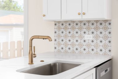 A gold kitchen faucet detail with white cabinets and marble countertop, gold hardware, and a mosaic tile backsplash.