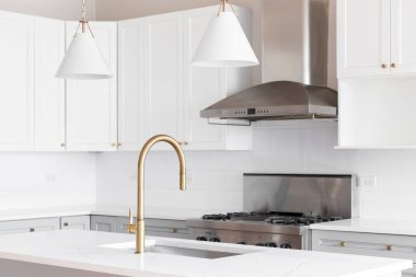 Beyaz ve gri dolaplı bir mutfak müfrezesi, beyaz mermer tezgahta altın musluk, adanın üzerinde asılı kolye ışıkları ve paslanmaz çelik aletler..