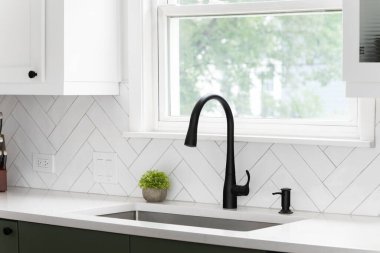 Lüks bir mutfakta beyaz ringa kemikli fayanslarla çekilen lavabo ayrıntıları. Beyaz mermer tezgah, beyaz ve yeşil dolaplar ve siyah bir musluk..