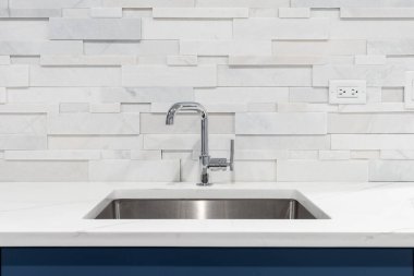 Modern bir mutfak lavabosu, beyaz mermer bir tezgahta krom musluk, mavi dolaplar, ve beyaz istiflenmiş taş bir arka plan..